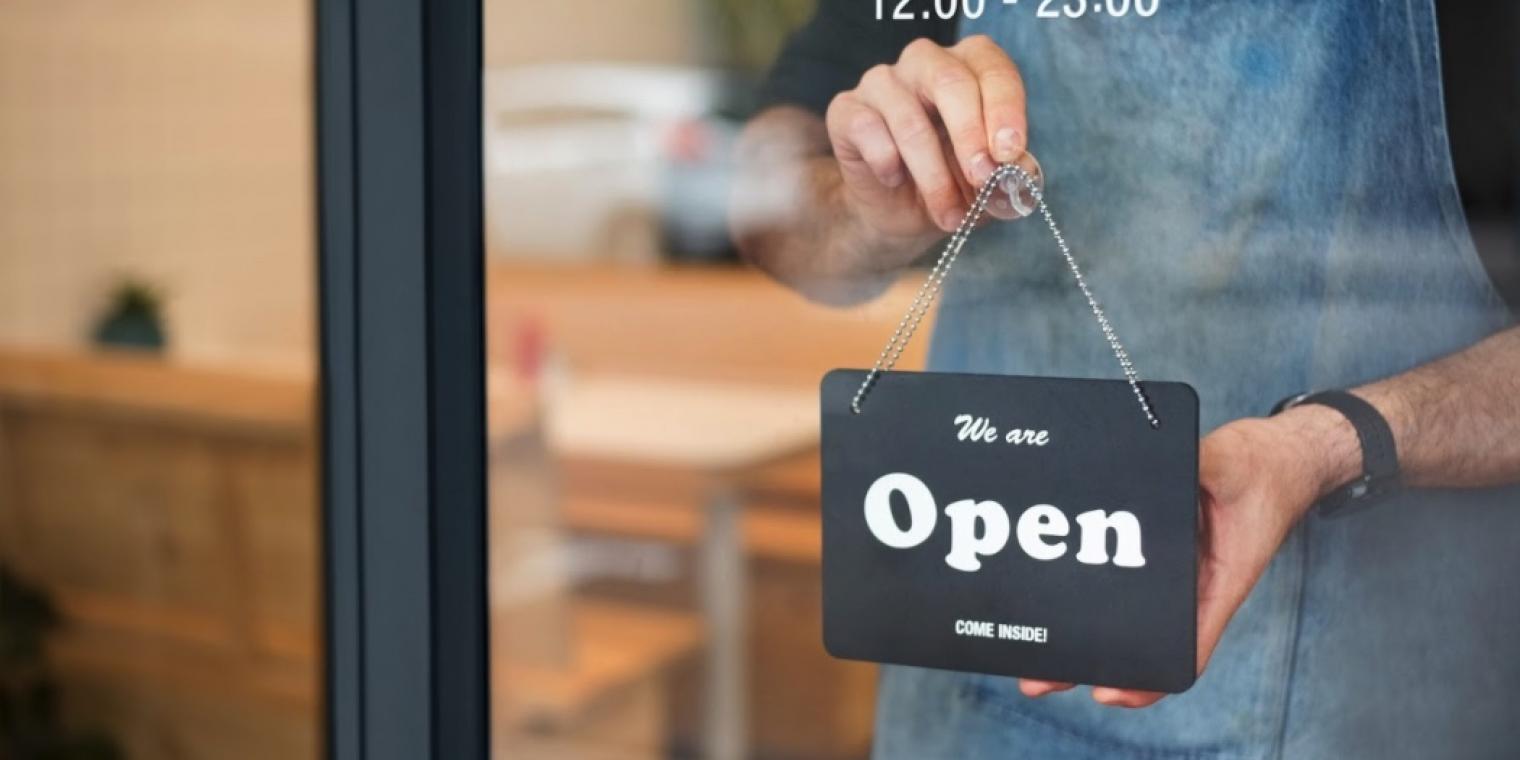A worker holds a sign that says "We are open, come inside!" at the entrance of a cafe.