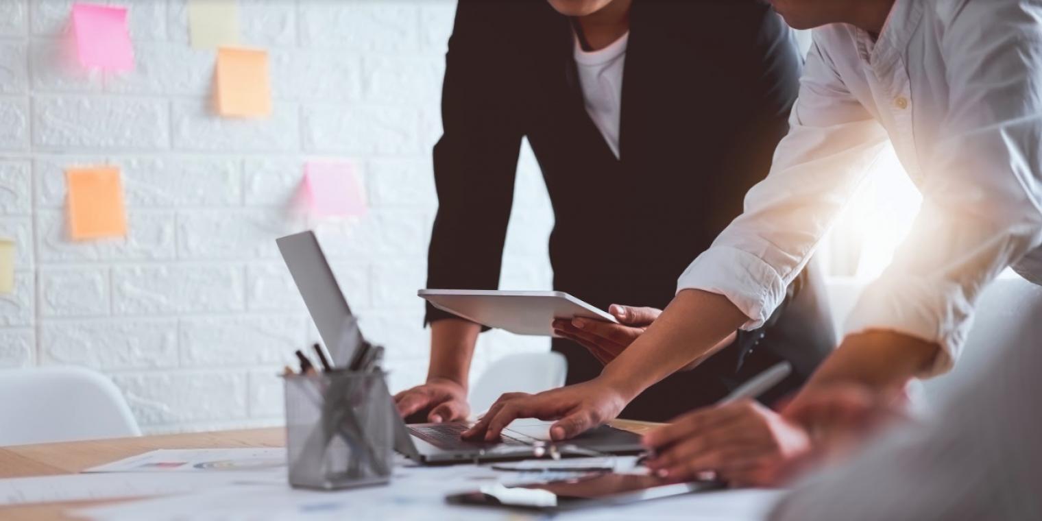 Two people stand over a laptop before a white wall with Post-It Notes on it.